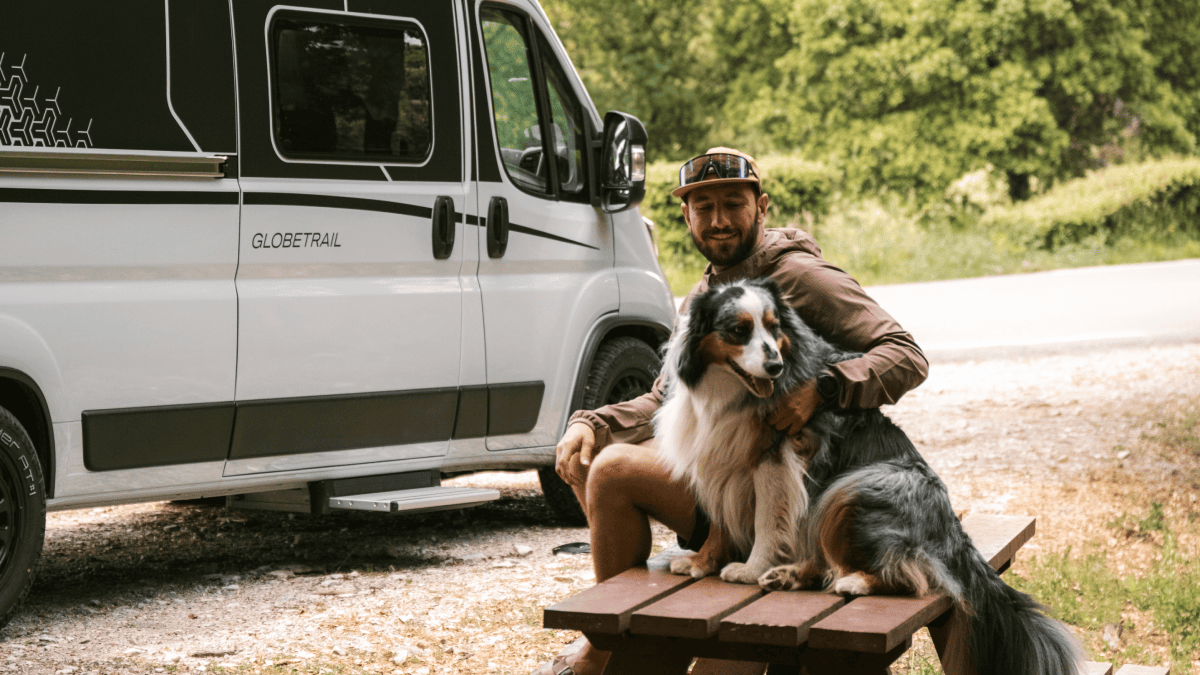 Man met hond op een bank naast een buscamper, ontspannen tijdens een actieve vakantie in de buitenlucht.